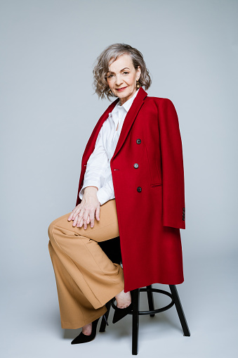 Elegance elderly lady wearing red coat and white shirt sitting on stool against grey background, smiling at camera. Studio shot of female designer.