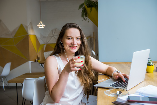 Portrait of smiling young business woman with coffee having a video call using laptop and headset sitting in cafe, coworking. Remote work, social networks, blogging. Telecommuting concept. Freelancer