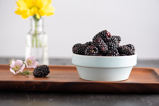 Fresh, ripe blackberries piled in a light blue bowl on a wooden serving tray. single blackberry off to the side with a flowering blackberry flower. Small vase with yellow flowers in the background.