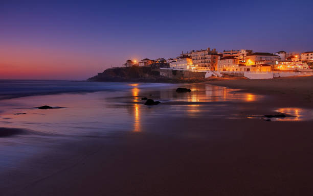 Amazing landscape of the Atlantic ocean coast at dusk. Night view on the village in lights, reflecting on a sandy beach. Long exposure image. Beach of Praia das Macas. Sintra. Portugal. stock photo