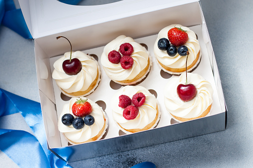 Cupcakes with whipped cream, fresh cherries, raspberries, blueberries, strawberries in gift box with blue ribbon. Selective focus.