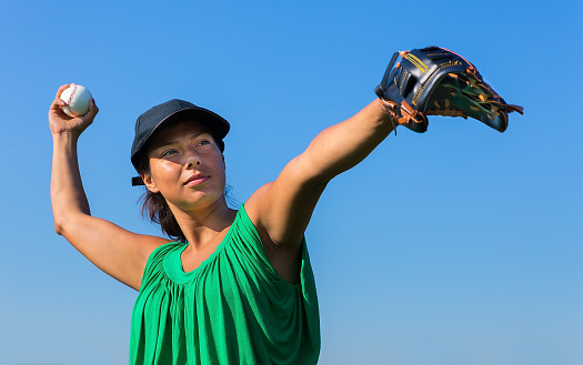 Colombian woman with baseball glove and cap throwing baseball in blue sky. On this sunny day in summer season the female person likes to practice sport outside. The woman is looking in the direction where she will be throwing.