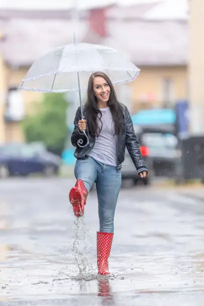 Happy woman in red polka dot rainboots  walks on the street with an umbrella on a raidy day.