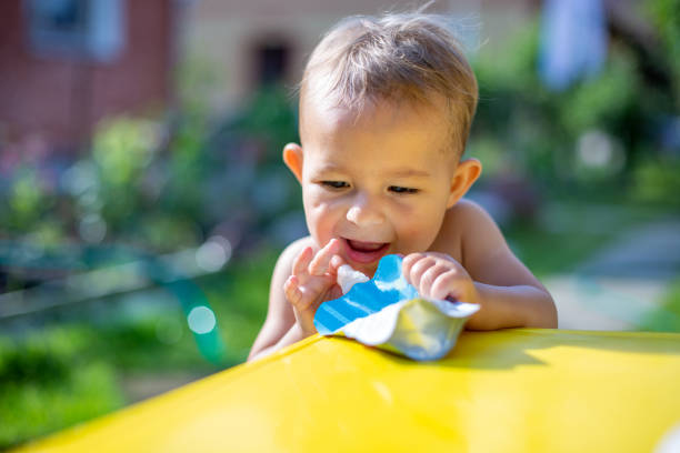 cute little baby put his finger in the package of fruit puree in pouch  in front of the yellow table. on the background is a green garden on a sunny day in blur, close up stock photo