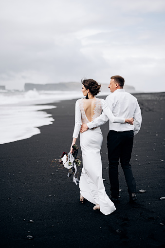 Destino boda islandia. Una pareja de bodas camina a lo largo de la playa negra de Vic. Sandy beach con arena negra en las orillas del Océano Atlántico. Enormes ondas espumosas. El novio abraza a la novia. photo