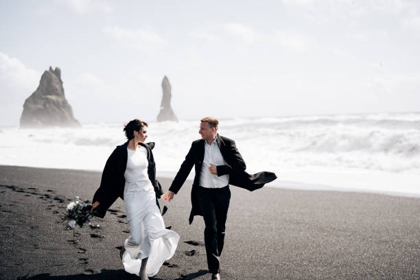 destination islande mariage. le couple de mariage court le long de la plage noire de sable de vik, près de la roche de basalte, sous la forme de piliers. se tenir la main, courir le long de la rive sur fond de vagues - elope photos et images de collection