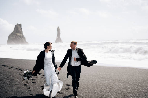 destinazione islanda matrimonio. la coppia nuziale corre lungo la spiaggia nera sabbiosa di vik, vicino alla roccia basaltica, sotto forma di pilastri. tenersi per mano, correndo lungo la riva sullo sfondo di onde - elope foto e immagini stock