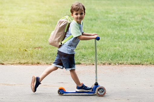 Smiling caucasian schoolboy running to school in a scooter carrying a backpack