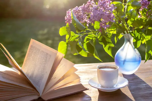 Photo of Little white cup of espresso coffee, opened book, blue semi-transparent vase with purple lilac flowers on rustic wooden table in the garden at spring morning after sunrise or at evening before sunset