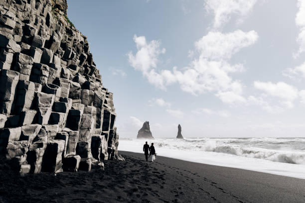 destination islande mariage. un couple de mariage marche le long de la plage noire de sable de vik, près de la roche de basalte, sous la forme de piliers. les mariés se tiennent la main et marchent le long de la rive. - elope photos et images de collection