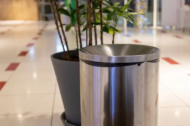 Photo of close-up steel cylinder bin litter bin. near in blur a plant in a gray tub. Against the background of a light floor tile and the interior of a public place in blur