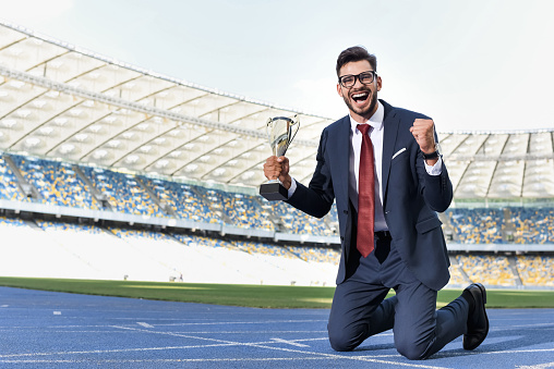 happy young businessman in suit standing on knees on running track with trophy at stadium