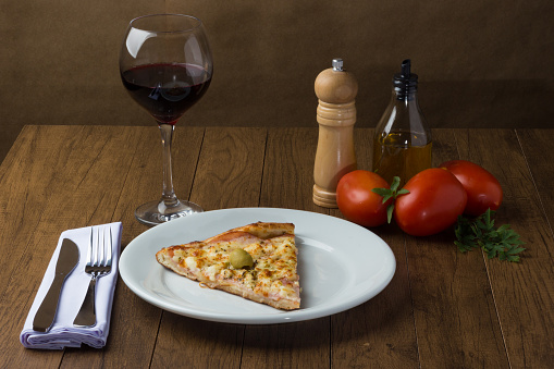 Gratin Loin Pizza. Mozzarella, Canadian Sirloin, Creamy Curd and Green Olives. Slice served on white dish. Wine Glass, Black Pepper Grinder, Basil, Olive Oil, Fork and Knife. Italian typical food.