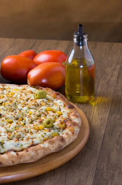 Photo of Pizza Chicken Catupiry flavor served on a wooden board. Made with Mozzarella, Chicken, Creamy Curd, Green Olives, Corn and Red Tomato Sauce.Olive oil and red tomatoes in the background.