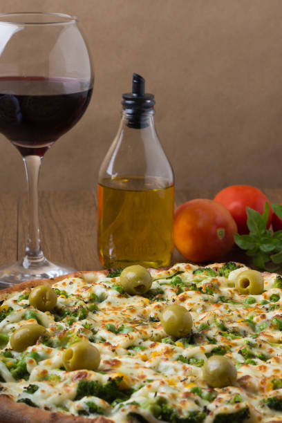 istock Broccoli pizza served on wooden board. Baked with mozzarella, broccoli, corn, peas, olives and sauce. Olive oil, basil, glass of red wine and tomatoes. vertical photo. 1240643521