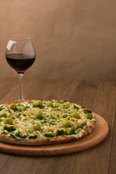 istock Delicious Broccoli Pizza served on wooden board. Baked with mozzarella, broccoli, corn, peas, olives and sauce. Olive oil, basil, glass of red wine. Vertical photo with great clarity. 1240643626