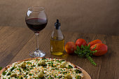 istock Delicious Broccoli Pizza served on wooden board. Baked with mozzarella, broccoli, corn, peas, olives and sauce. Olive oil, basil, glass of red wine and tomatoes. Pizza on the left. Featured in wine. 1240644157