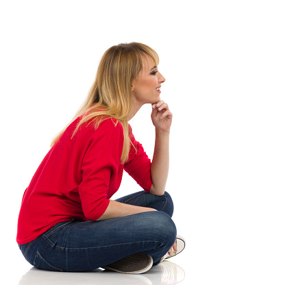 Young woman in red sweater sits on a floor, holds hand on chin, looks away and smiles. Side view. Full length studio shot isolated on white.