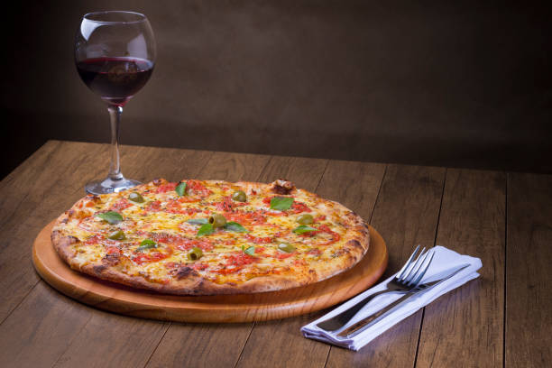 pizza marguerita made with tasty pizza dough, mozzarella, tomatoes, marjoram and green olives. served on a wooden board. napolitan pizza. red wine served in a crystal glass. fork and knife on a napkin - crystal noodles imagens e fotografias de stock