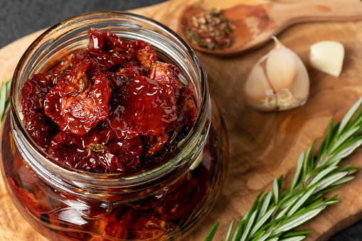 Sun-dried tomatoes with garlic, rosemary and spices in a glass jar on an olive wood cutting board.