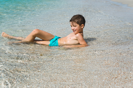 A cheerful kid on the beach on a summer sunny day lies in the clear sea water and looks happy.
