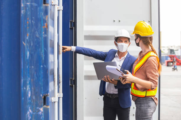 homme et femme de contremaître de travail utilisant le masque de protection et le casque de sécurité utilisant l’ordinateur portatif et retenant des récipients de vérification de presse-papiers dans le cargo pour l’exportation d’importation, con - inspecteur photos et images de collection