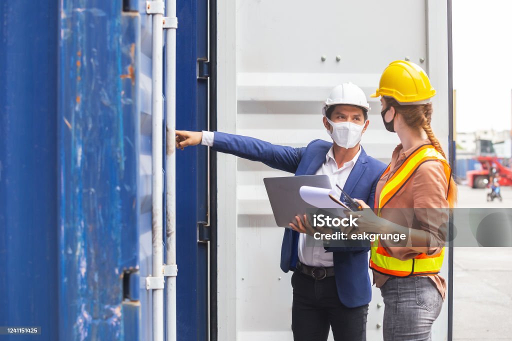 Teamwork foreman man and woman wearing protection face mask and safety helmet using laptop and holding clipboard checking containers in cargo ship for import export, Industrial container cargo concept. Construction Industry Stock Photo