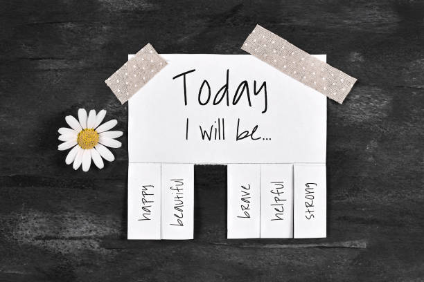 White tear-off stub note with text 'Today I will be...' and words 'happy, beautiful, brave, helpful' and 'strong' and wone stub missing on dark black background with blooming cute daisy flower