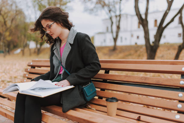 Pretty woman sitting with coffee abd book outdoors Beautiful female student wearing glasses and leafing through pages of book in a park while studying urban dictionary stock pictures, royalty-free photos & images