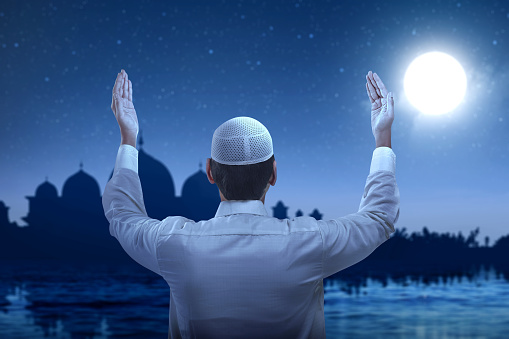 Rear view of Asian Muslim man standing while raised hands and praying with the night scene background
