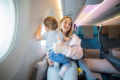 concept everything is OK. lofe is good. mom shows thumb up and looks at the camera with a smile. little cute toddler jumping on her knees with a young beautiful mother in an airplane chair.