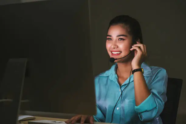 Photo of Asian Female customer care service smiling and working hard late in night shift at office, call center department, worker and overtime, teamwork with colleagues for success concept