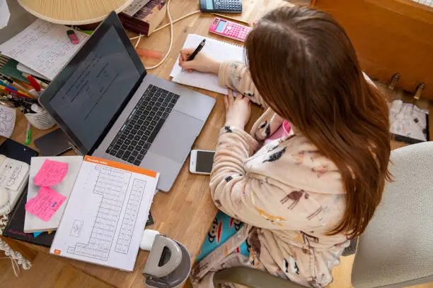 A teenage Caucasian girl sits at a desk at home studying. She is using a laptop and pen with note book.