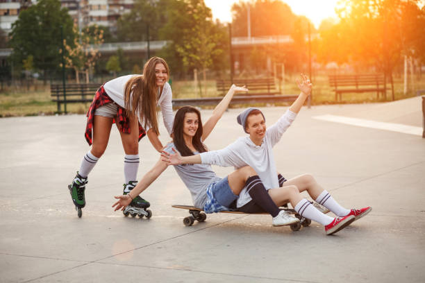 Full length shot of three happy teenage girl having fun at the skate part. One girl on the roller blades is pushing the skateboard with two of her friends sitting on it with their arms in the air. They are all smiling and looking at camera.