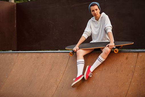 Full length shot of smiling teenage girl sitting on a sports ramp and taking a break from riding her skateboard that she is holding.