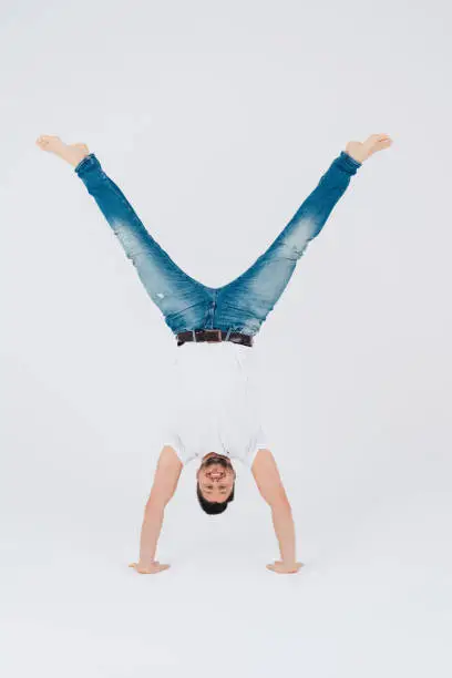 Athletic smiling barefoot young man in jeans performing a handstand with wide open legs over a white studio background