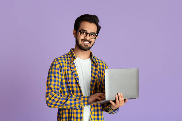 Smiling Indian guy with laptop computer working or studying online on lilac background Happy Indian guy with laptop computer working or studying online on lilac background indian young man stock pictures, royalty-free photos & images