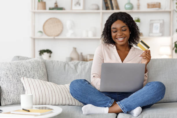 compras en línea. chica negra positiva usando el ordenador portátil y la tarjeta de crédito en casa - online shopping fotografías e imágenes de stock
