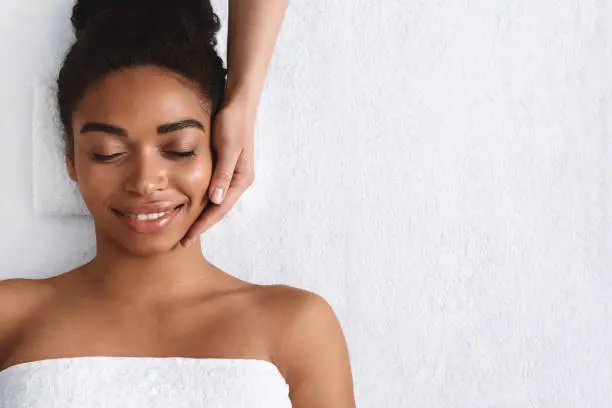 Joyful african girl with closed eyes having face massage at spa, top view, copy space