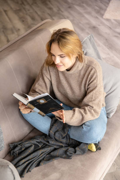 istock beautiful young girl reads a book while sitting on a sofa, bending legs 1241365879