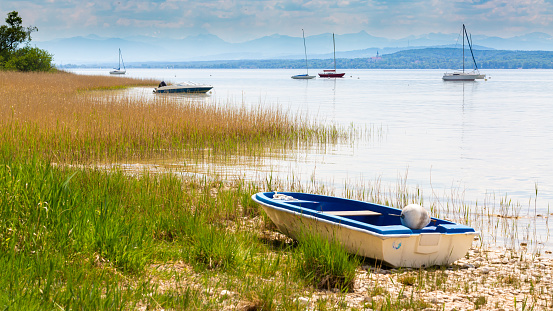 Stegen, Bavaria / Germany - May 19, 2020: Rowing boat on the shore of Ammersee (Lake Ammer). Sailboats and the silhouette of the alps in the background. Beautiful bavarian landscape. Panorama format.