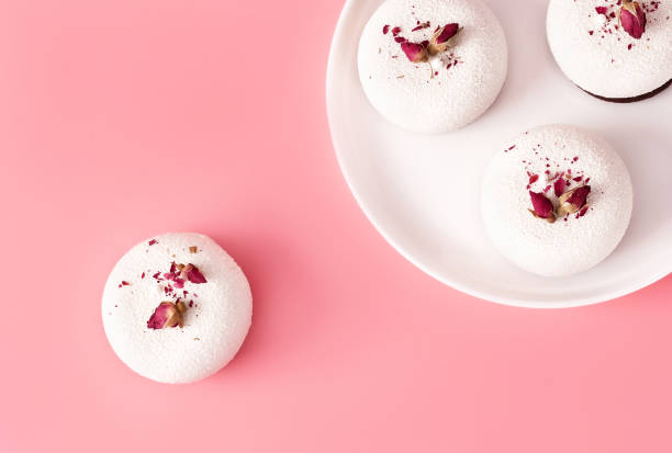 istock Mousse pastry dessert covered with white chocolate decorated with rose buds on pastel pink background. Modern stylish cake. Flat lay style. 1241431162