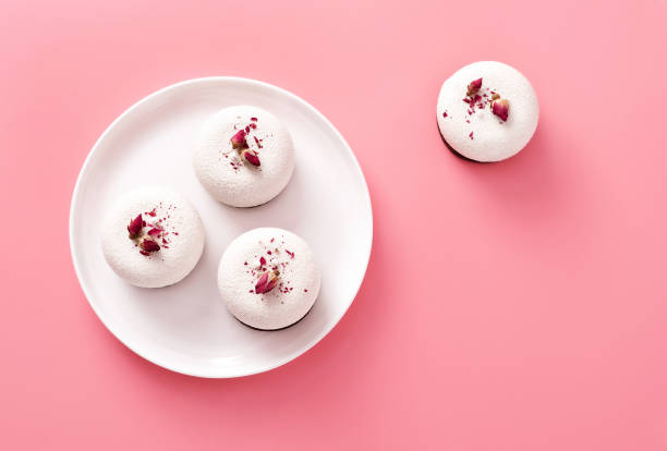 istock Mousse pastry dessert covered with white chocolate and decorated with rose buds on pastel pink background. Modern stylish cake. Flat lay style. Copy space. 1241431164
