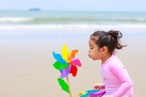 A little cute girl having fun on sandy summer with blue sea, playing on tropical beach with colorful paper windmill