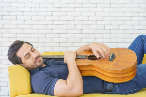 hombre feliz tocando la guitarra mientras está sentado en el sofá en la sala de estar, disfrutando del tiempo despreocupado en casa. - 16713 fotografías e imágenes de stock