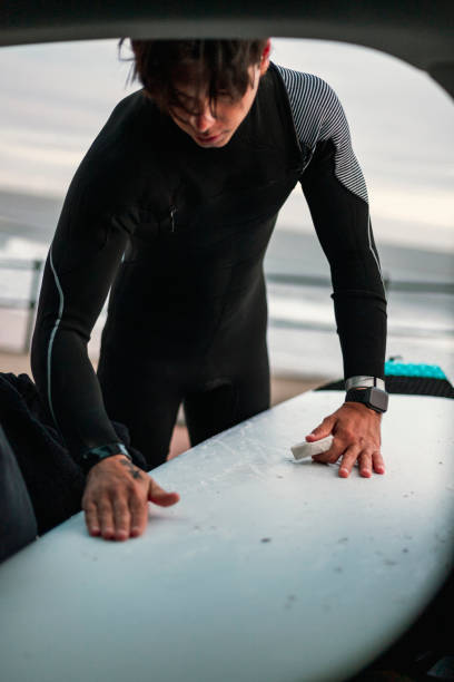 A young Caucasian man wearing a wetsuit, waxing his surfboard, preparing to go surfing at the beach. The shot has been taken from inside a car, where the surfboard is placed while the man stands outdoors.