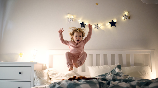 Two small laughing children jumping on bed indoors at home, having fun.