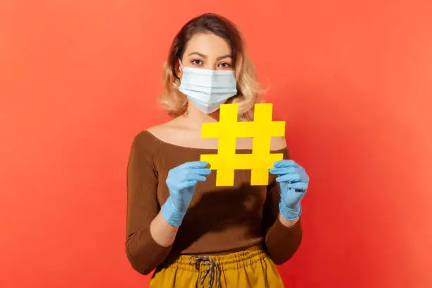 Photo of Tagged message Stay home. Woman wearing hygienic face mask gloves and holding hashtag symbol