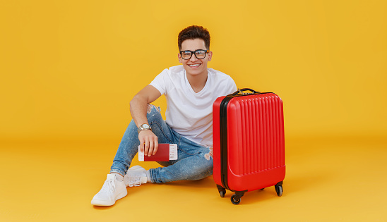 Ethnic young man with luggage smiling and looking at camera on yellow backdrop before summer vacation