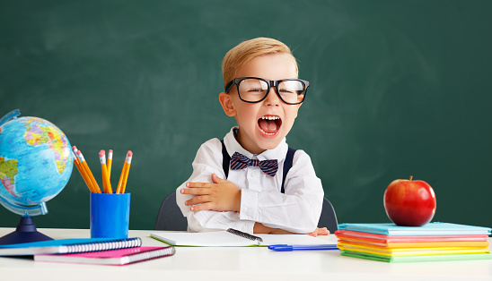 funny happy child   schoolboy    student sitting at   table and laughs near school blackboard